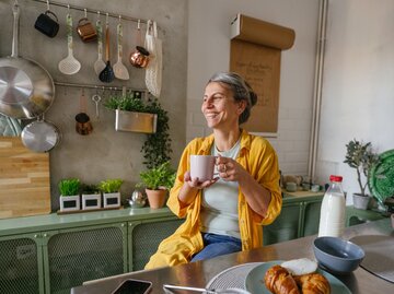 Eine Frau sitzt am Frühstückstisch mit Kaffee, Milch und Croissant  | © Getty Images / AleksandarNakic