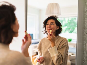 Eine Frau mit braunen kurzen Haaren trägt Lippenstift vor dem Spiegel auf | © Getty Images / Westend61