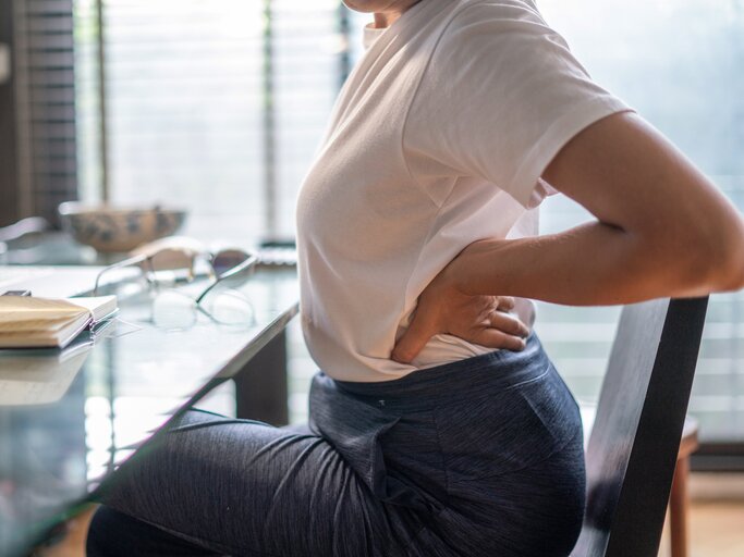 Frau sitzt am Schreibtisch und hält sich den Rücken | © GettyImages/	boonchai wedmakawand