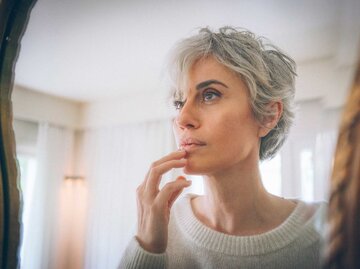 Frau mit kurzen grauen Haaren steht vor einem Spiegel und betrachtet ihr Gesicht. | © Getty Images / Ekin Ozbicer