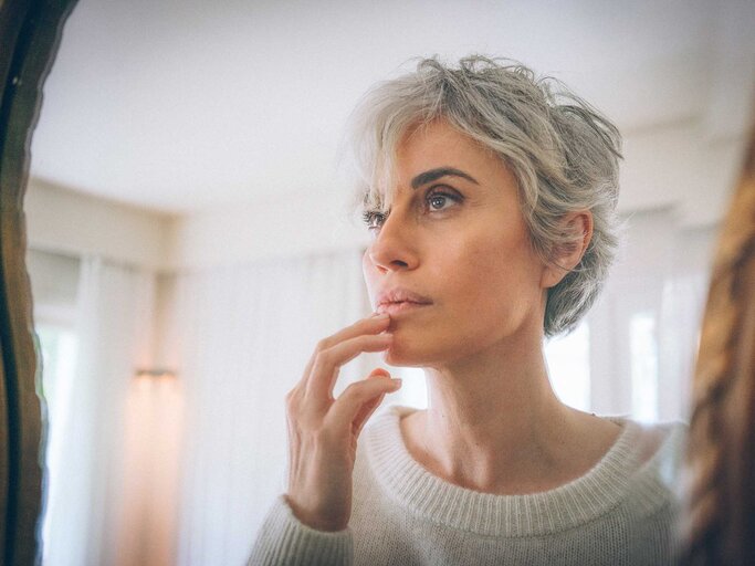 Frau mit kurzen grauen Haaren steht vor einem Spiegel und betrachtet ihr Gesicht. | © Getty Images / Ekin Ozbicer