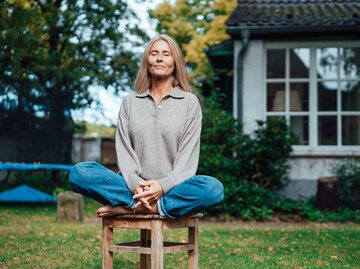 Frau sitzt im Garten auf einem Hocker und meditiert. | © Getty Images / Westend61
