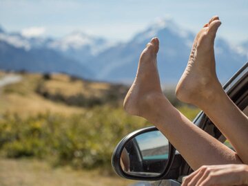 Frau hält vor einem Bergpanorama die Füße aus dem Autofenster | © Getty Images/swissmediavision