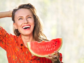 Frau mit langen, braunen Haaren und einer roten Bluse isst lachend eine Melone | © Getty Images/Peppersmint