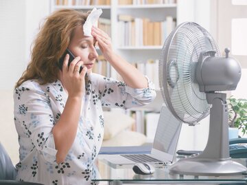 Frau sitzt vor Ventilator und wischt sich Schweiß von der Stirn | © Getty Images/humonia