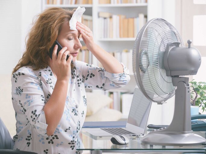 Frau sitzt vor Ventilator und wischt sich Schweiß von der Stirn | © Getty Images/humonia