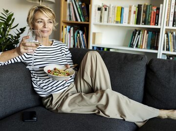 Frau sitzt mit Nudelgericht und einem Glas Wein auf dem Sofa | © Getty Images/Westend61
