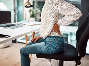 Frau sitzt an Schreibtisch hält sich Rücken | © Getty Images/PeopleImages