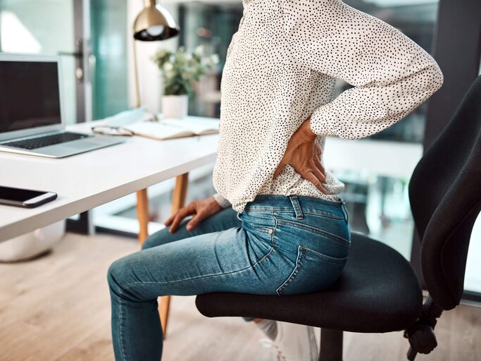Frau sitzt an Schreibtisch hält sich Rücken | © Getty Images/PeopleImages
