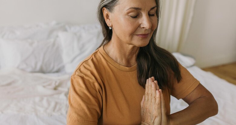 Frau mit langen, grauen Haaren und gelbem T-Shirt sitzt auf Bett und meditiert | © AdobeStock/shurkin_son