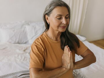 Frau mit langen, grauen Haaren und gelbem T-Shirt sitzt auf Bett und meditiert | © AdobeStock/shurkin_son