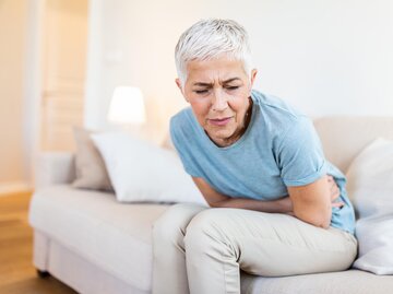 Frau mit kurzen, grauen Haaren sitzt auf Sofa und hält sich schmerzenden Bauch | © AdobeStock/Graphicroyalty