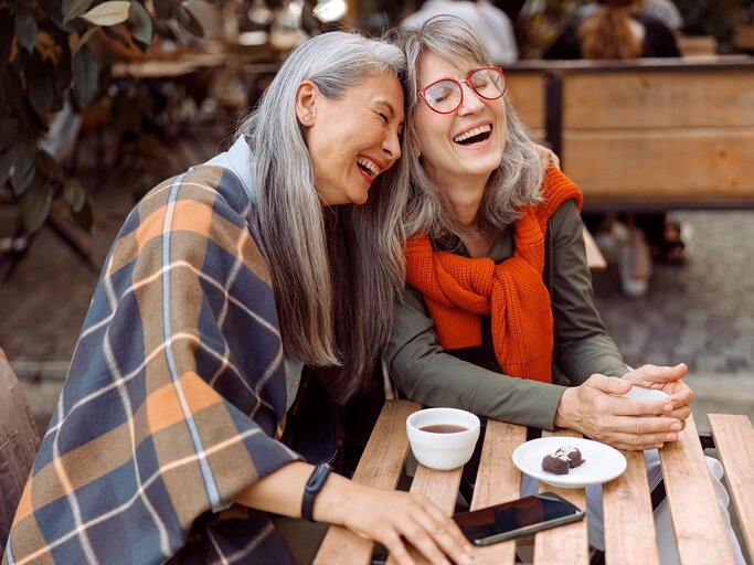 Zwei Frauen mit grauen Haaren sitzen an einem Tisch und lachen  | © AdobeStock/Yaroslav Astakhov