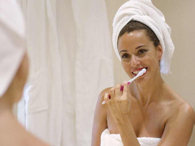 Frau steht mit Handtuch auf dem Kopf vor dem Badezimmerspiegel und putzt sich die Zähne. | © Adobe Stock/Designpics
