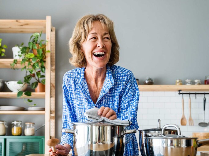 Frau steht in der Küche am Herd und hält den Deckel eines Topfes. | © Adobe Stock/oneinchpunch
