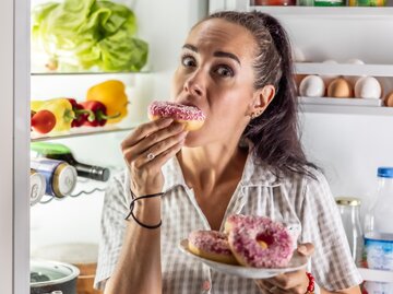 Frau steht vor offenem Kühlschrank und isst Donuts | © AdobeStock/weyo