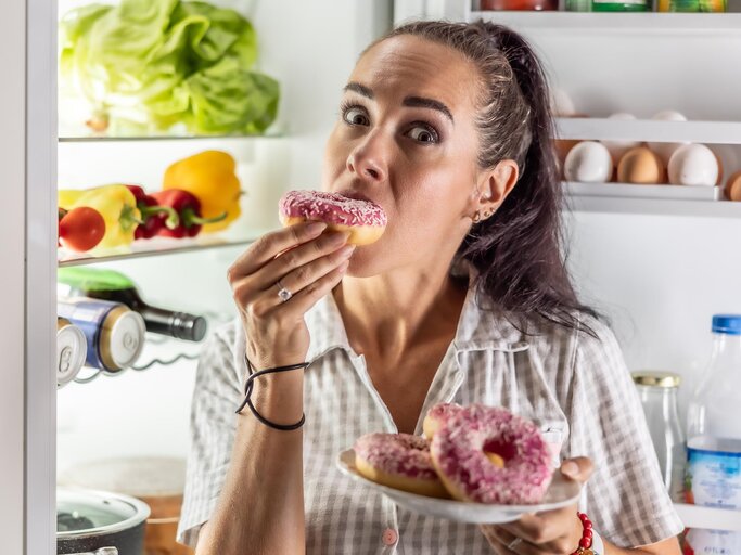 Frau steht vor offenem Kühlschrank und isst Donuts | © AdobeStock/weyo