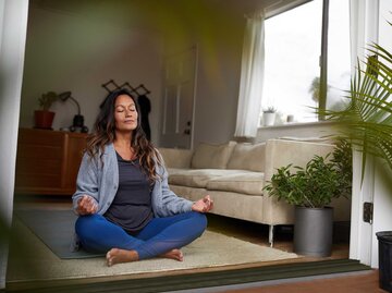 Reife Frau mit langen dunklen Haaren sitzt im Wohnzimmer auf dem Teppich und meditiert. | © Getty Images / Goodboy Picture Company