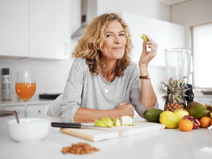 Ältere Frau steht in der Küche und isst einen Apfel | © Getty Images/PeopleImages