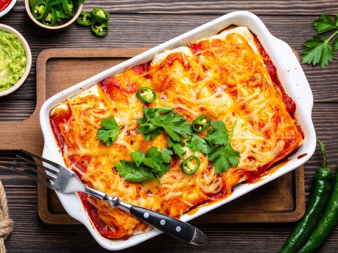 Enchiladas mit Fleisch, chiliroter Sauce und Käse in weißer Auflaufform über rustikalem Holzhintergrund, serviert mit Guacamole und Tomatendips | © Getty Images/thesomegirl