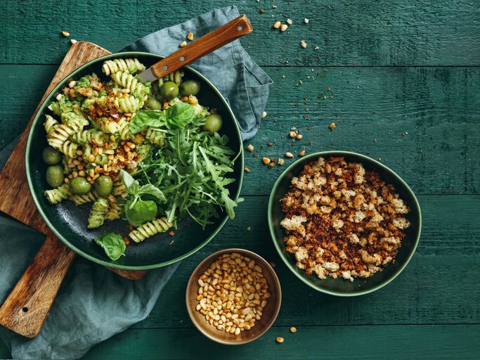 Sommerlicher vegetarischer Nudelsalat mit Brokkolipesto, Erbsen, Rucola, Oliven, Pinienkernen und Semmelbröseln auf dunkelgrünem Hintergrund. Draufsicht. | © Getty Images/luchezar