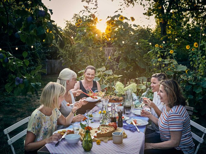 Freundinnen sitzen bei Sonnenuntergang um einen Tisch im Garten | © Getty Images/Uwe Krejci