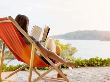 Person in Liegestuhl an See mit Buch und Weinglas | © Getty Images/Morsa Images