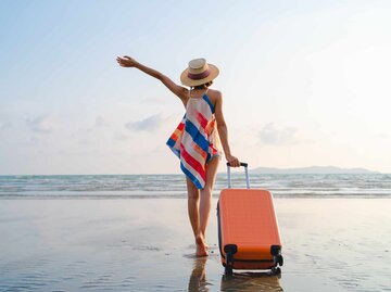 Glückliche Frau in Urlaubsstimmung mit Rollkoffer am Strand | © Getty Images/Twenty47studio