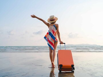 Glückliche Frau in Urlaubsstimmung mit Rollkoffer am Strand | © Getty Images/Twenty47studio