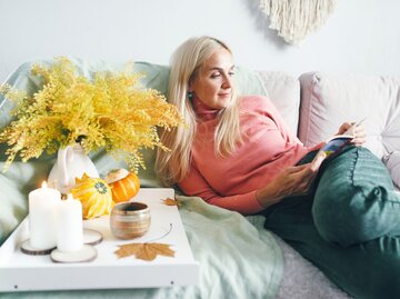 Portrait einer Frau, die mit Herbstdeko auf der Couch liegt und liest  | © Getty Images / Ekaterina Goncharova