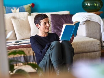 Frau mit kurzen Haaren sitzt im Wohnzimmer auf dem Boden vor der Couch und liest ein Buch. | © Getty Images / Oliver Rossi