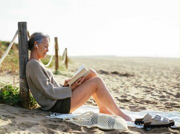 Frau mit grauen Haaren, braunem Pullover und schwarzen Shorts liest ein Buch am Strand | © Getty Images/Westend61