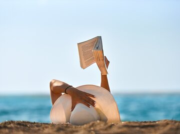 Eine Frau liest am Strand ein Buch | © GettyImages/NDinfinity