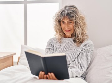 Frau sitzt im Bett und liest Buch | © AdobeStock/Krakenimages.com