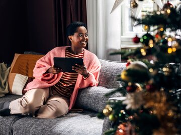 Eine Frau sitzt auf der Couch und liest. Neben ihr steht ein geschmückter Weihnachtsbaum. | © Getty Images / FreshSplash