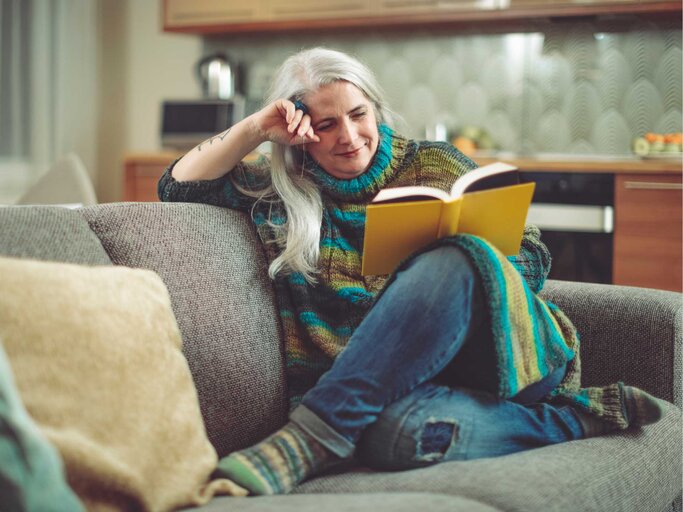 Frau in mittlerem Alter sitzt auf der Couch und liest ein Buch | © Getty Images/Eva-Katalin