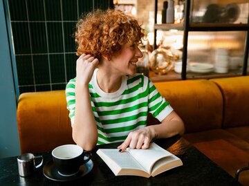 Frau mit roten Locken und gestreiftem T-Shirt sitzt im Café und liest ein Buch | © AdobeStock/Drobot Dean