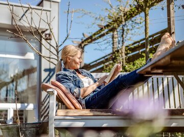 Eine Frau sitzt auf einer sonnigen Terrasse und liest | © Getty Images / Westend61