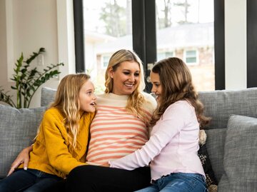 Frau sitzt mit ihren beiden Töchtern lächelnd auf der Couch und unterhält sich | © Getty Images/MoMo Productions