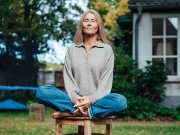 Frau sitzt im Scheidersitz auf einem Hocker im Garten und hat die Augen geschlossen | © Getty Images/Westend61