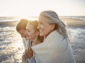Drei Generationen von Frauen halten sich am Strand beim Sonnenuntergang in den Armen. | © Getty Images / Oliver Rossi