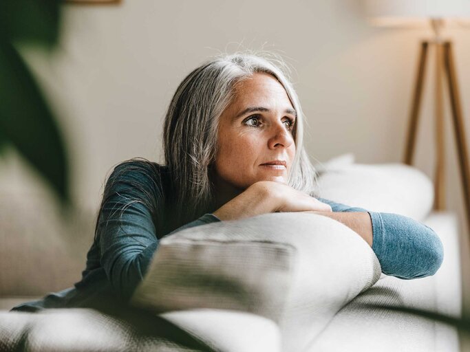 Frau mit grauen langen Haaren sitzt auf der Couch und schaut nachdenklich. | © Getty Images / Westend61
