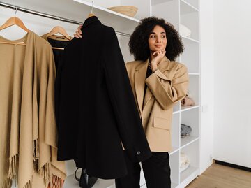 Frau in beigem Blazer und schwarzer Hose hält schwarzen Blazer in der Hand und überlegt | © Getty Images/DeanDrobot