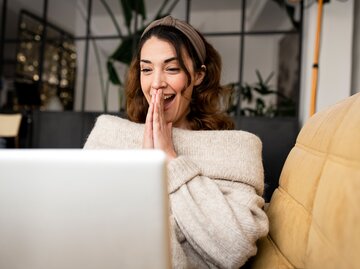 Eine Frau freut sich und sitzt vor ihrem Laptop auf einem gelben Sofa | © GettyImages/Cavan Images