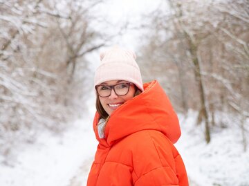 Frau mit oranger Daunenjacke steht in verschneiter Landschaft | © AdobeStock/sepy