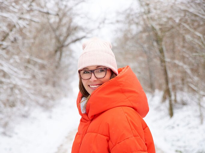 Frau mit oranger Daunenjacke steht in verschneiter Landschaft | © AdobeStock/sepy