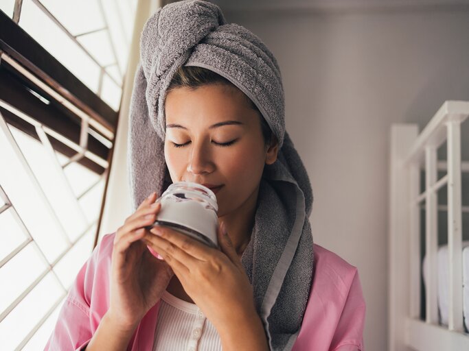 Eine Frau riecht an einem Glas voll Kokosöl und hat ein Handtuch auf dem Kopf | © GettyImages/FreshSplash