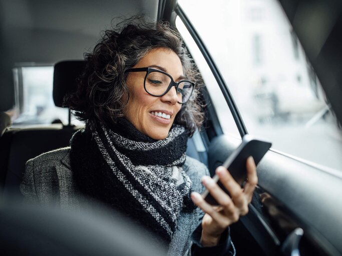 Frau mit Brille im Auto sieht lächelnd auf ihr Handy | © gettyimages.de | Luis Alvarez