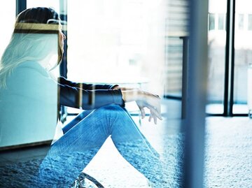 Frau mit Burnout sitzt mit angezogenen Knien auf dem Boden hinter einer Glasscheibe. | © Tinpixels, Getty Images