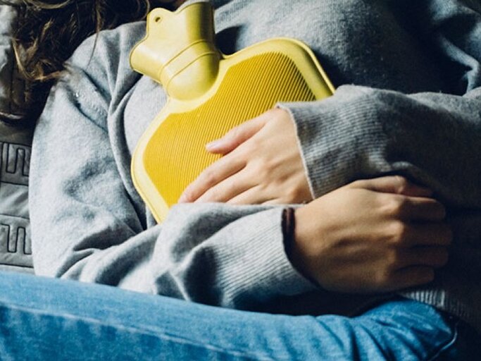 Frau liegt mit gelber Wärmflasche auf dem Bauch im Bett. | © Carlo107, Getty Images